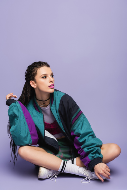 woman with braids sitting in nineties style jacket and looking away on purple background - Foto, Bild