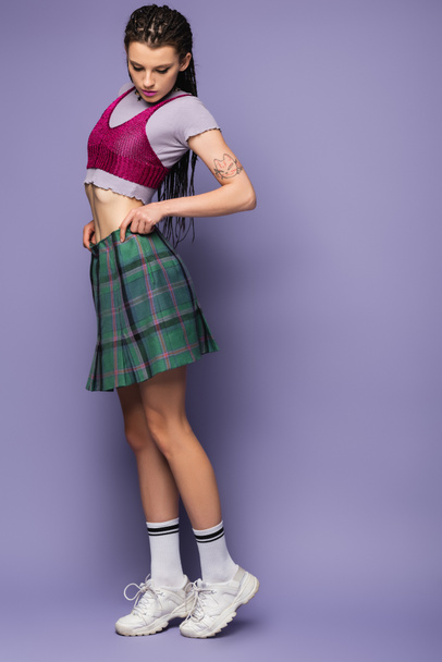 full length of woman in crop top and white sneakers adjusting checkered skirt on purple background - Foto, Imagem