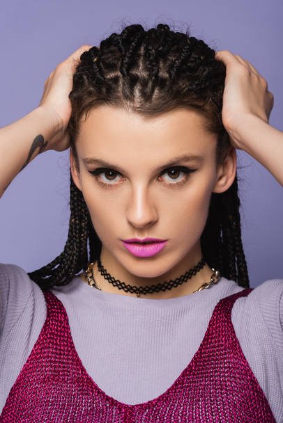 portrait of pretty woman in makeup touching braided hair while looking at camera isolated on purple - Foto, immagini