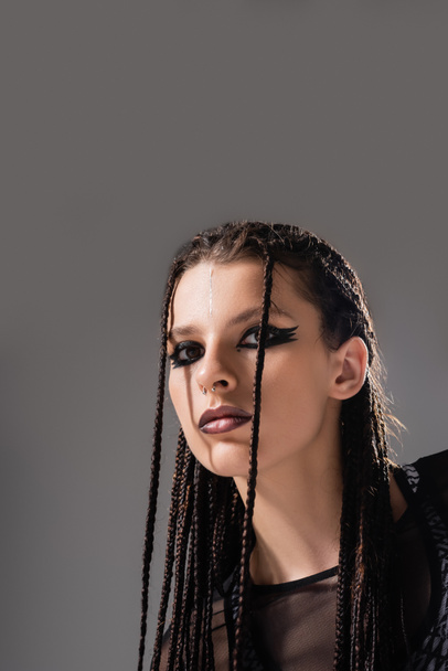 portrait of young woman with futuristic makeup and braided dreadlocks looking at camera isolated on grey - Foto, Imagen
