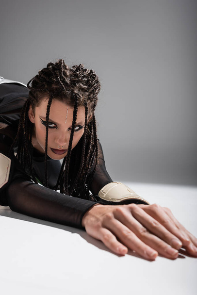brunette woman with futuristic makeup and dreadlocks posing on white surface and grey background - Fotoğraf, Görsel