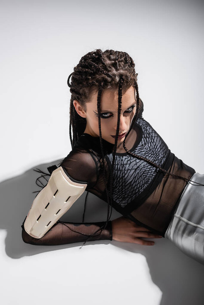 brunette woman with braids and expressive makeup lying in futuristic outfit on grey background - Photo, Image
