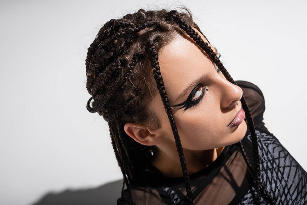 portrait of young woman with braids and futuristic makeup looking away on grey background - Foto, Imagen
