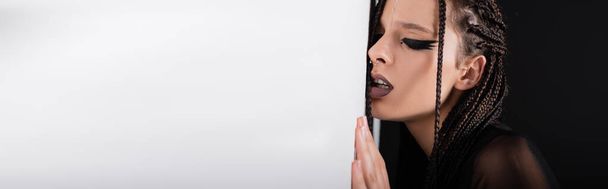 експресивна жінка з футуристичним макіяжем і косами, спираючись на стіну на чорному тлі з білим копіювальним простором, банер
 - Фото, зображення