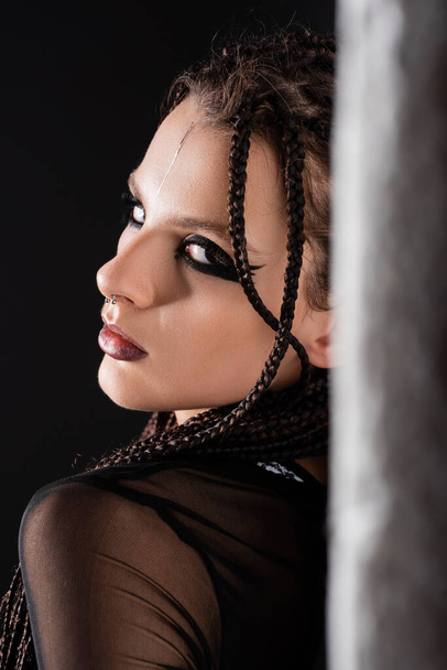 portrait of woman with dreadlocks and stylish makeup looking at camera near blurred wall on black background - Photo, Image
