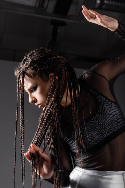 futuristic woman in black outfit touching braided dreadlocks while posing on grey background - Photo, Image