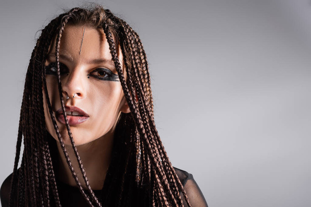 portrait of woman with braids and stylish makeup with piercing looking at camera isolated on grey - Photo, Image