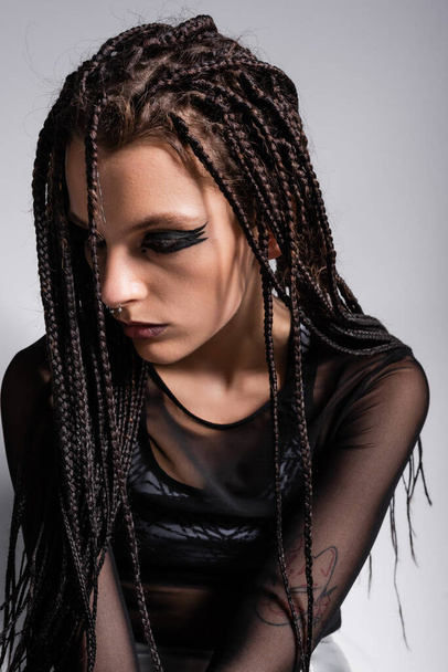 brunette woman with braids posing in futuristic makeup and black clothes isolated on grey - Foto, Imagem
