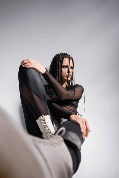 trendy woman with dreadlocks sitting in black futuristic outfit on grey background - Foto, imagen