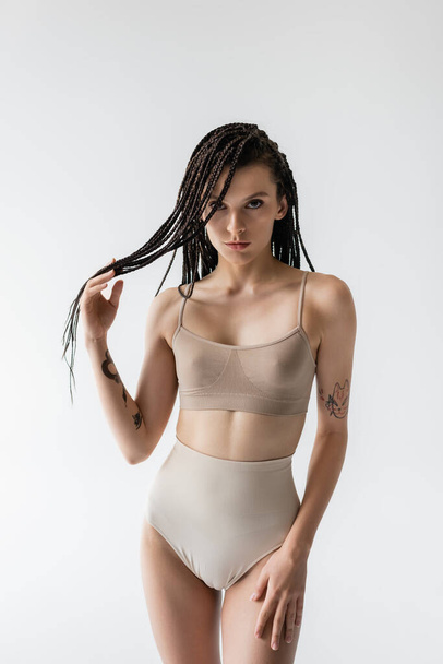 tattooed woman with perfect body touching braids and looking at camera isolated on grey - Photo, Image
