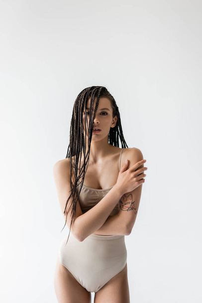 brunette woman with braided dreadlocks hugging herself while posing in lingerie isolated on grey - Photo, Image