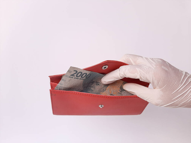 A hand in white latex glove holds a red purse with money 2000 IDR, Indonesian banknotes. Isolated in white background. Side view. - Foto, afbeelding