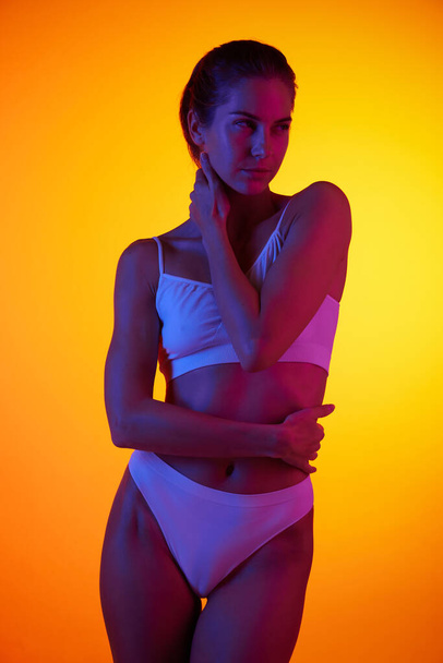 Portrait of beautiful woman posing in underwear isolated over orange studio background in neon light. Self care. Concept of beauty, body and skin care, health, plastic surgery, cosmetics - Fotografie, Obrázek