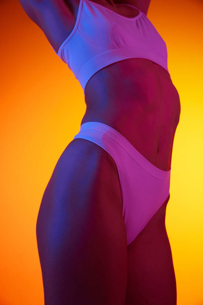 Snímek štíhlého ženského těla v bílém spodním prádle izolovaném na oranžovém pozadí v neonovém světle. Fitness životní styl. Koncept krásy, péče o tělo a pokožku, zdraví, plastická chirurgie, kosmetika - Fotografie, Obrázek