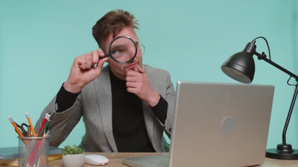 Tutkija tutkija parrakas liikemies istuu toimistotiskillä pitäen suurennuslasia lähellä kasvoja, katsoen kameraan, jolla on iso zoomattu hauska silmä, etsien, analysoiden sinisellä taustalla - Materiaali, video