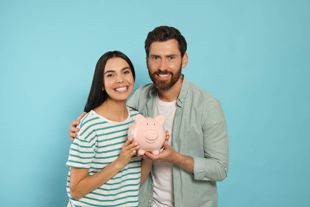 Happy couple with ceramic piggy bank on light blue background - Foto, Imagen