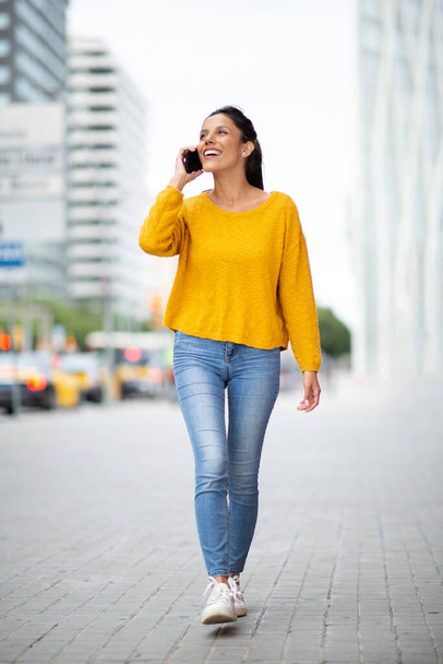 Full body portrait smiling woman walking and talking with cellphone in city  - Foto, Bild