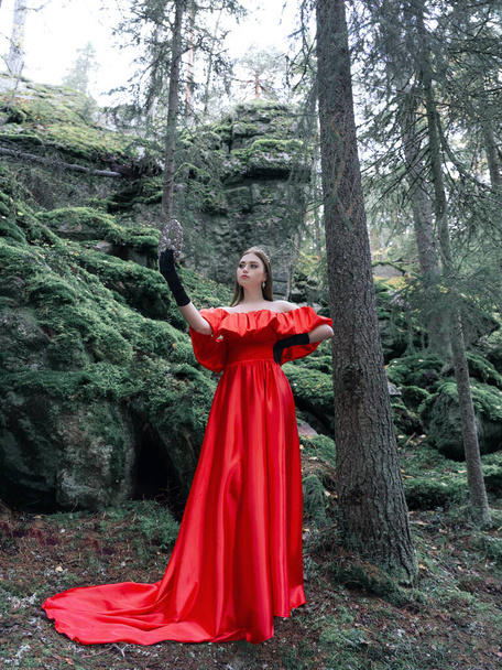 Een mooie vrouw in een lange rode jurk, zwarte satijnen handschoenen en een diadeem op haar hoofd tegen de achtergrond van een bos. De bosnimf kijkt in de spiegel. Volledig portret. - Foto, afbeelding
