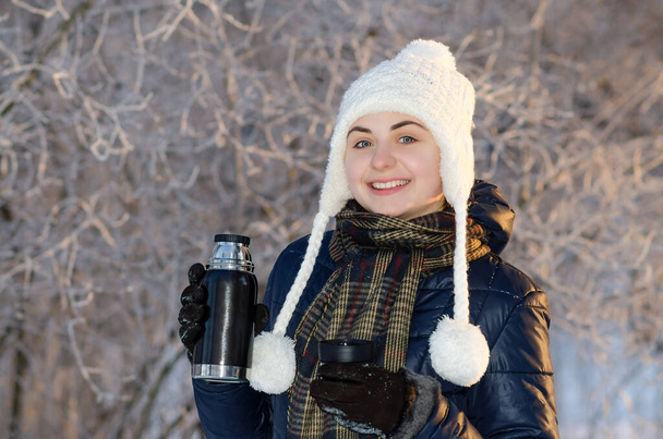 Boldog fiatal nő mosolyog és egy termoszt tart a havas téli parkban meleg kötött sapkában. Napos idő. Téli szezon. - Fotó, kép