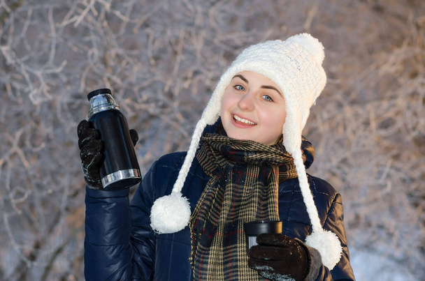 Gelukkige jonge vrouw lachend en een thermosfles vasthoudend in besneeuwd winterpark met warme gebreide hoed. Zonnig weer. Winterseizoen. - Foto, afbeelding
