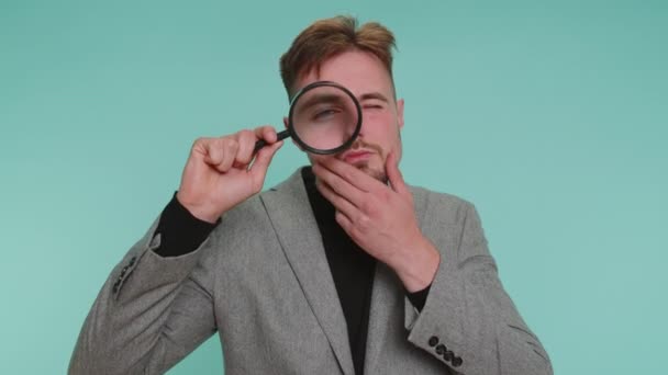 Investigator researcher scientist business man holding magnifying glass near face, looking into camera with big zoomed funny eyes, searching, analysing. Young guy isolated on blue studio background - Footage, Video