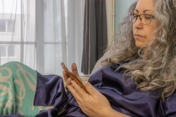 Idősebb nő kényelmesen fekszik az ágyban mobiltelefonnal a kezében, nyugodt arckifejezéssel, híreket kap, lábadozik a betegség után, ősz-fekete hosszú hullámos haj, szemüveg, pizsama. Wellness koncepció otthon - Fotó, kép