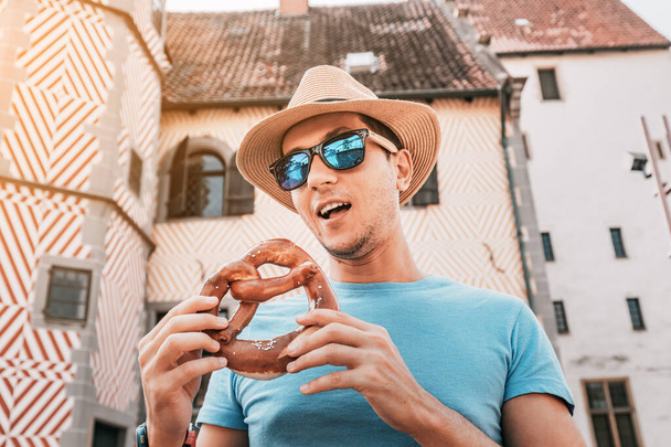 Gelukkig mannelijk toerist eten heerlijke pretzel snack op de achtergrond van een oud Duits gebouw. Reizen en toerisme keuken - Foto, afbeelding