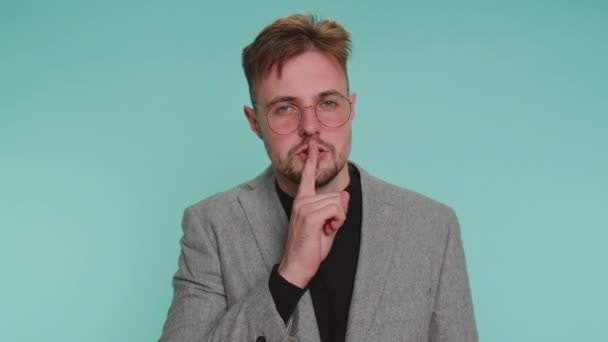 Shh be quiet please. Bearded adult business man 20 years old presses index finger to lips makes silence gesture sign do not tells secret. Young adult handsome guy posing on blue studio background - Footage, Video