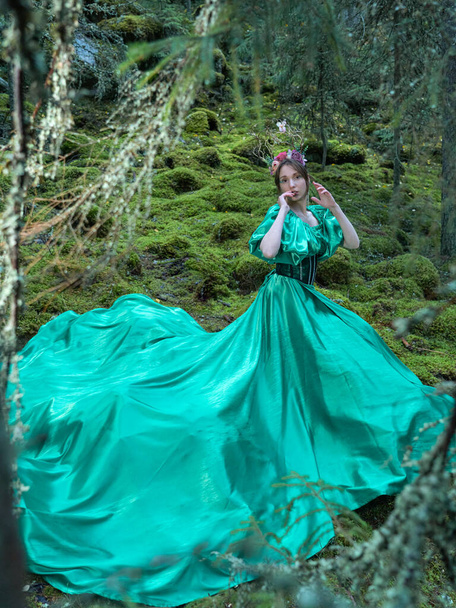 Een mooie vrouw in een lange groene jurk met een korset en een decoratieve kroon van takken en bloemen op haar hoofd. Bosnimf. Volledig portret. - Foto, afbeelding