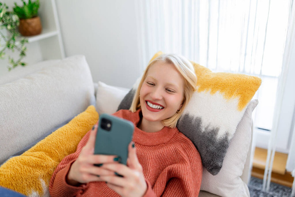 Young woman sit relax on sofa in living room browsing surfing wireless internet on smartphone, millennial girl rest on couch at home message text on modern cellphone, shopping online via website - Valokuva, kuva
