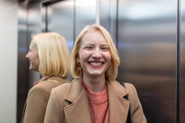 Schattige blonde vrouw in trendy jas verblijf in lift alleen en glimlachend, vrouw in lift - Foto, afbeelding