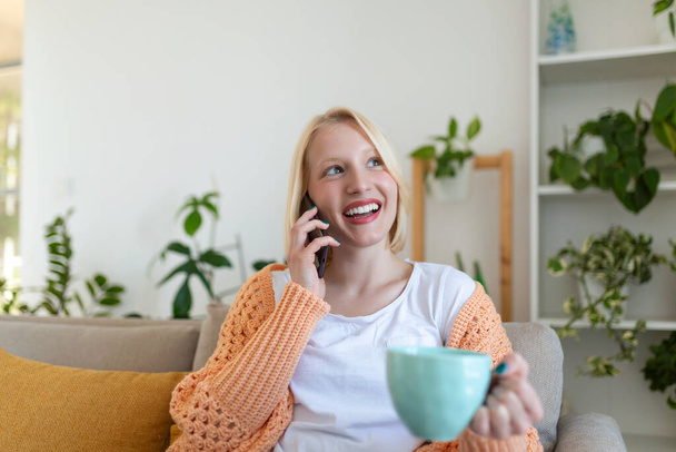 Houkutteleva hymyilevä nainen, joka käyttää älypuhelinta istuessaan sohvalla kotona. Viestintä ja kodikkuus käsite
. - Valokuva, kuva