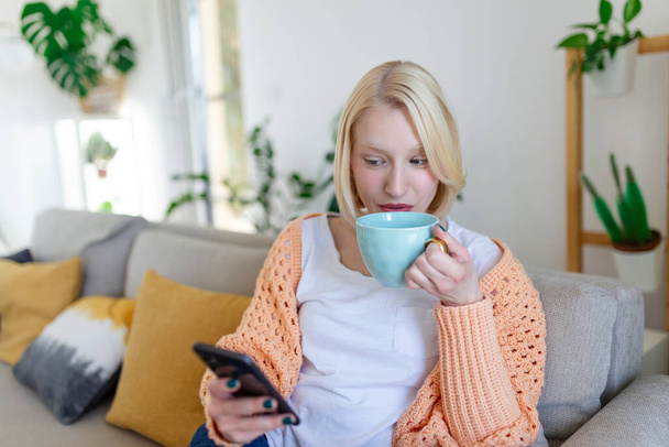 Attractive smiling woman using smart phone while sitting on the sofa at home. Communication and coziness concept. - Foto, Bild