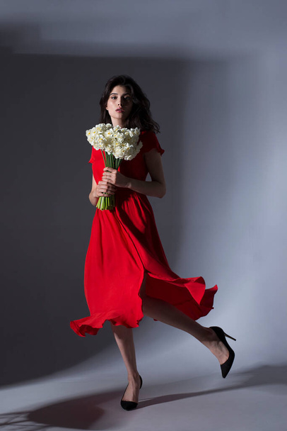 Full length image of a brunette woman in a red dress holding a bouquet of white daffodils and spinning, against a gray background. - Photo, Image