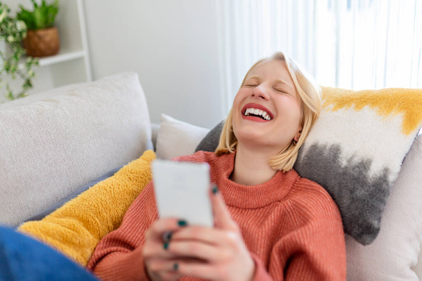 Νεαρή γυναίκα κάθεται να χαλαρώσετε στον καναπέ στο σαλόνι περιήγηση surfing ασύρματο internet στο smartphone, χιλιετή κορίτσι ξεκουραστούν στον καναπέ στο σπίτι μήνυμα κειμένου στο σύγχρονο κινητό τηλέφωνο, ψώνια σε απευθείας σύνδεση μέσω της ιστοσελίδας - Φωτογραφία, εικόνα