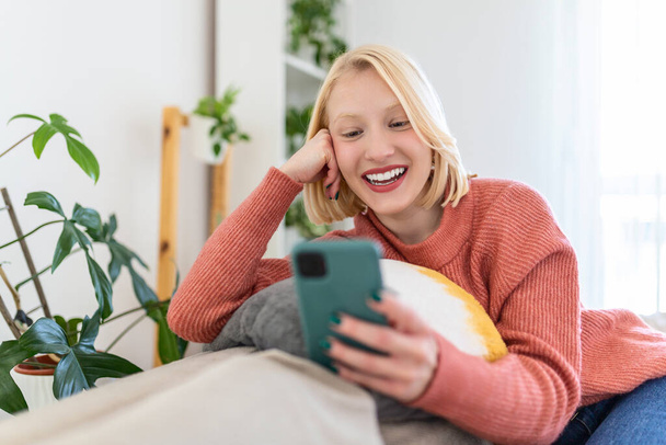 Atractiva mujer sonriente usando un teléfono inteligente mientras está sentada en el sofá de su casa. Concepto de comunicación y comodidad
. - Foto, imagen