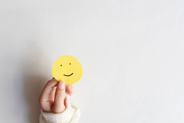 Vrouw handen houden gele glimlach gezicht papier op witte achtergrond, bovenaanzicht, goede feedback rating, positief denken, klantbeoordeling, beoordeling, wereld geestelijke gezondheid dag, Compliment Day. - Foto, afbeelding