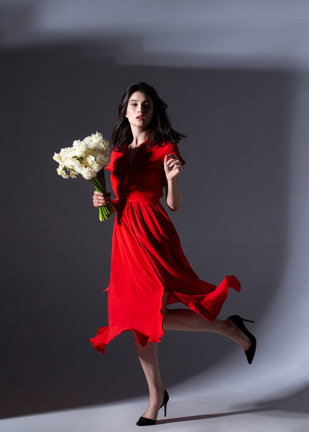 Full length image of young sexy woman posing in a red dress holding a bouquet of white daffodils, over grey background. - Photo, Image