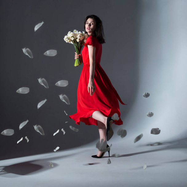 Teljes hosszúságú kép fiatal elegáns gyönyörű nő pózol egy piros ruha gazdaság egy csokor fehér nárcisz, felett szürke háttér. - Fotó, kép