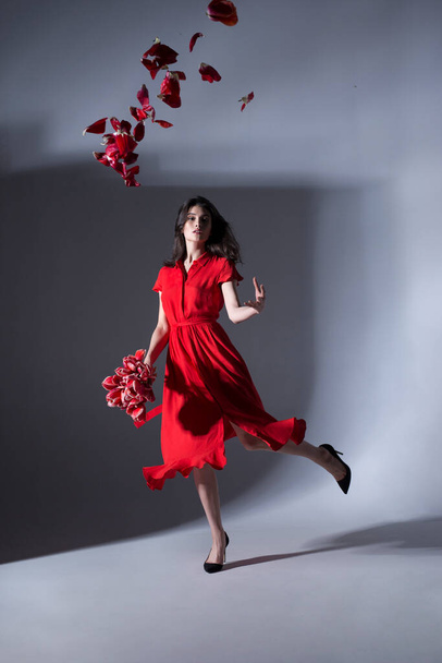 Full length image of young elegant woman posing in a red dress holding a bouquet of white daffodils, over grey background. - Fotografie, Obrázek