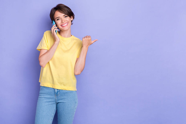 Portrait photo of young adorable cute lady wear bright yellow t-shirt speak phone toothy smile speak finger point empty space isolated on purple color background. - Фото, изображение