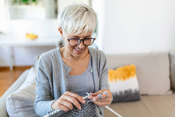 Starší žena v brýlích sedět na gauči doma úsměv drží pletací jehly a nitě pleteniny oblečení pro milované, oblíbená činnost a zábava, v důchodu klidný bezstarostný život koncept - Fotografie, Obrázek