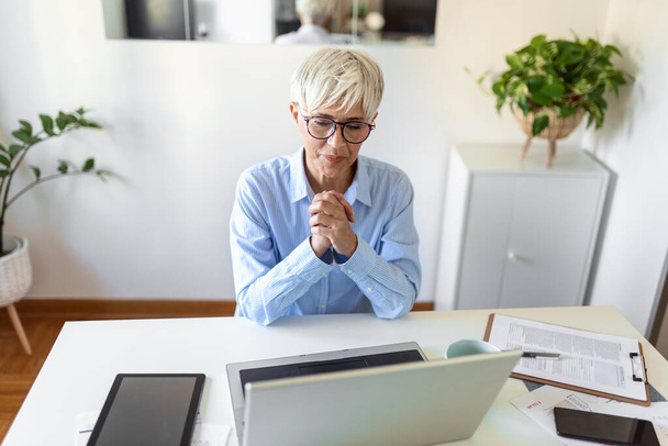 Πορτρέτο της casual γυναίκα χρησιμοποιώντας το laptop της, ενώ κάθεται στο σπίτι γραφείο και εργασίας. Μια ελκυστική μεσήλικη επιχειρηματίας κάθεται μπροστά από το φορητό υπολογιστή και τη διαχείριση της μικρής επιχείρησής της από το σπίτι. - Φωτογραφία, εικόνα