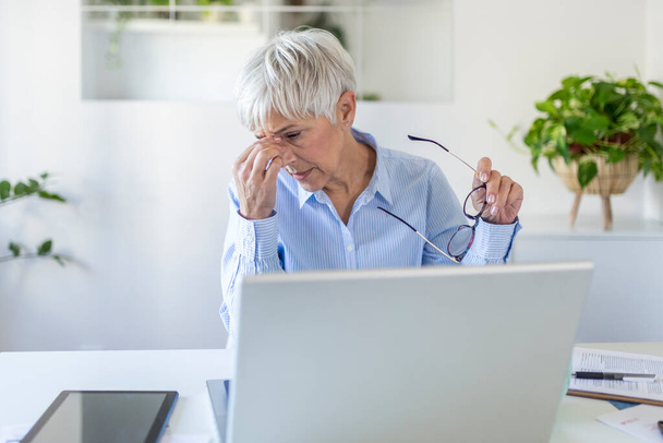 Mujer mayor frustrada que trabaja desde la oficina en casa delante de la computadora portátil, con dolor de cabeza. Mujer madura que tiene una expresión dolorosa de la cara - Foto, Imagen