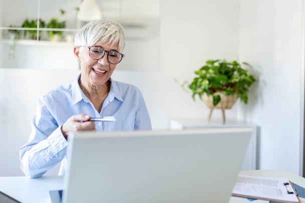 Kobieta nosi okulary w domu koncentrując jak ona działa na laptopie. Kobieta w średnim wieku pracuje z domu na laptopie. Portret wykonawczej dojrzałej kobiety wpisując na laptopie podczas pracy w domu. - Zdjęcie, obraz