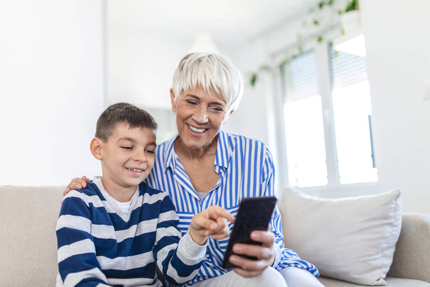 Abuela sosteniendo a su nieto en la habitación de casa. Una mujer mayor con un niño mirando el teléfono. Feliz abuela con su nieto en casa. - Foto, imagen