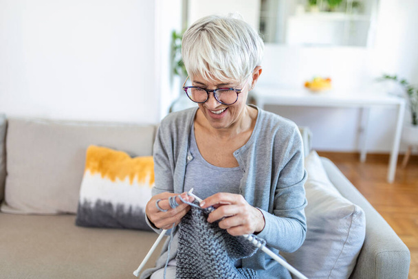 眼鏡をかけた年配の女性は、家庭の笑顔でソファに座り、編み物の針と糸のニットを愛する人のための服、好きな活動と娯楽、引退した静かなのんきな生活の概念 - 写真・画像