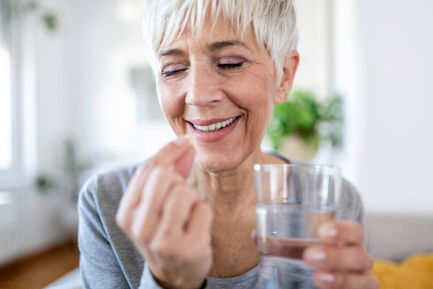 Smiling happy healthy middle aged 50s woman holding glass of water taking dietary supplement vitamin pill. Old women multivitamins antioxidants for anti age beauty. - Фото, изображение