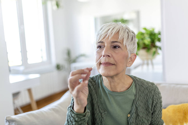 woman using cotton swab while doing coronavirus PCR test at home. Woman using coronavirus rapid diagnostic test. Mature woman at home using a nasal swab for COVID-19. - Photo, Image
