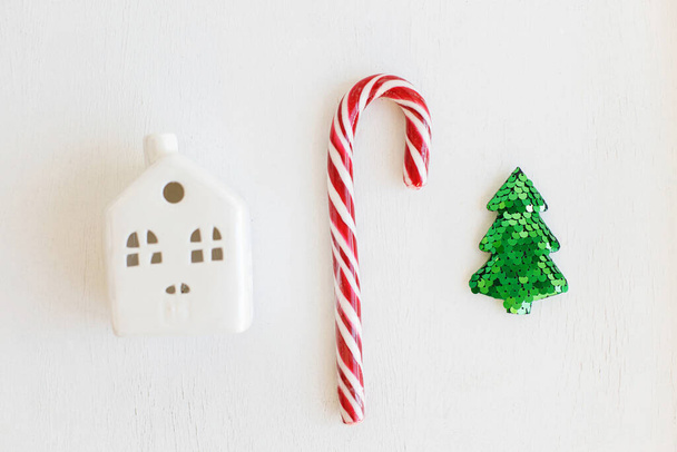メリークリスマスとハッピーホリデー!白いテーブルの上にスタイリッシュな小さなクリスマスツリー、家やお菓子の杖とクリスマスフラットレイ組成物。現代のお祝いの冬のバナー、スキャンディ装飾 - 写真・画像